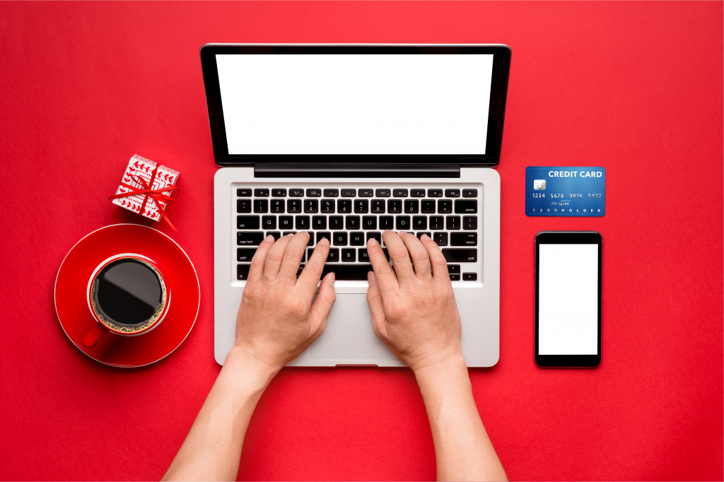 A person rests their hands on their laptop keys with a cellphone, credit card, cup of coffee and small wrapped box around it.